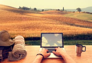 Digital nomads are people who can work from anywhere using a laptop and an internet connection: from an hillside italian town in Tuscany a young male traveller is typing on his laptop computer on a wooden table, with a travel backpack and a mug with coffee. On the background a beautiful panorama of hills covered by corn fields and trees at sunset. Laptop display reflects sky with clouds.